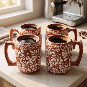 May include: Four terracotta-colored ceramic mugs filled with dark coffee, each featuring a white hand-painted floral and swirl pattern. The mugs have sturdy handles and a textured rim detail, arranged in a two-by-two grid on a light countertop.
