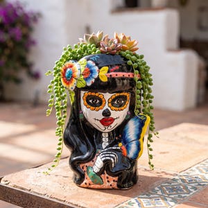 May include: A black ceramic planter shaped like a Day of the Dead figure with a white face, red lips, and floral accents. The figure features long black hair, a blue butterfly on the shoulder, and is filled with green string of pearls and succulents.