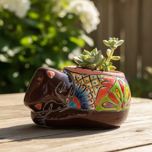 May include: A brown ceramic planter in the shape of a sleeping animal, with a colorful floral design on the side. The planter holds a small succulent plant. The planter is sitting on a wooden surface.