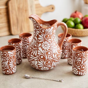 May include: A terracotta pitcher and six matching tumblers featuring a white hand-painted floral and swirl pattern. The set includes a single small silver-toned metal spoon placed in front of the pitcher on a flat surface.