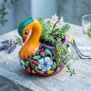 May include: A ceramic swan-shaped planter featuring a glossy orange head and neck with a green crown. The body is black with a hand-painted floral pattern in blue, white, green, and orange. It contains a small succulent plant with tiny white flowers.
