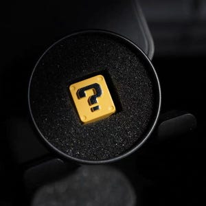 May include: A yellow square with a black question mark, set in a black circular container. The container is surrounded by a dark background. The design is reminiscent of a video game.