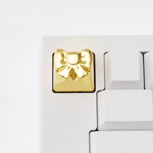 May include: A gold-colored bow-shaped keycap on a white keyboard. The keycap has a square base and a detailed bow design. The keyboard keys are white, creating a contrast with the gold keycap.