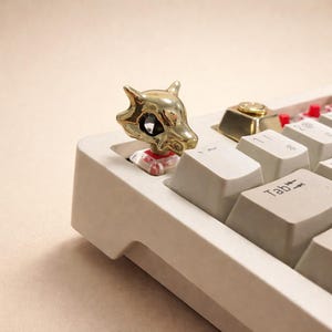 May include: A gold-colored, stylized skull keycap on a mechanical keyboard. The keycap is a detailed, three-dimensional design. The keyboard has off-white keys and a gold accent key. The keycap is a unique accessory for computer enthusiasts.