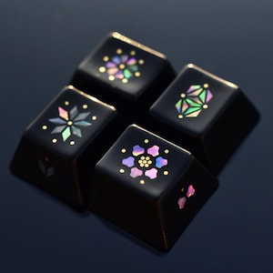 May include: Four black keyboard keycaps with iridescent floral and geometric designs. Each keycap features a different pattern, including flowers and starbursts, with gold accents. The keycaps are arranged in a square formation against a dark background.