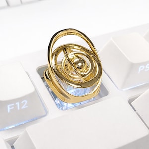 May include: A gold-colored, decorative ring stand in the shape of an armillary sphere, resting on a white keyboard key. The stand features concentric rings and a central sphere, with a loop for holding. The keyboard keys are white.