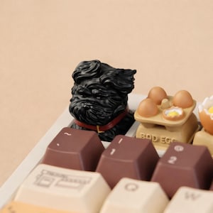 May include: A black dog figurine with a red collar sits atop a keyboard. The keyboard keys are various colors, including brown and cream. A small egg-shaped figurine with the text "BAD EGGS" is also visible.