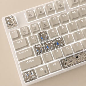 May include: A close-up of a white mechanical keyboard with custom keycaps. The keycaps have a unique design with silver and blue accents. The keyboard has standard keys, including function keys, number keys, and a shift key.
