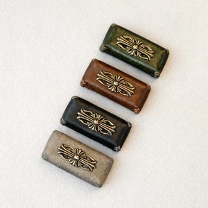 Può includere: Quattro scatole rettangolari in pelle verde, marrone, nera e grigia, ciascuna con un emblema decorativo in metallo dorato. Le scatole sono disposte diagonalmente su una superficie bianca, mostrando la varietà di colori e design.