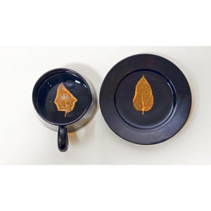 May include: A black teacup and saucer set with a gold leaf design. The teacup is filled with a dark liquid, and the saucer has a single leaf in the center. The set is on a white surface.