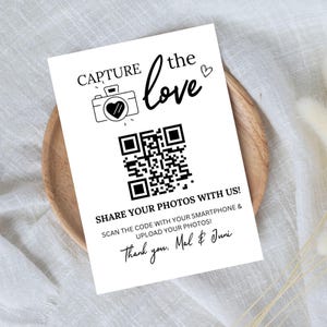 May include: Black and white wedding sign with a QR code, a camera icon with a heart, and the text "CAPTURE the love" and "SHARE YOUR PHOTOS WITH US! SCAN THE CODE WITH YOUR SMARTPHONE & UPLOAD YOUR PHOTOS! Thank you, Mal & Juni"