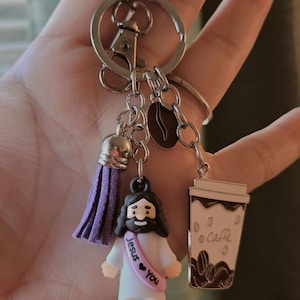 May include: A silver keychain with multiple charms. Charms include a purple tassel, a coffee bean, a coffee cup with the word "Caffe", and a small Jesus figurine with a sash that says "Jesus &hearts; You".