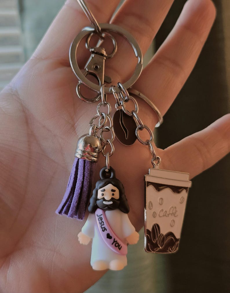 May include: A silver keychain with multiple charms. Charms include a purple tassel, a coffee bean, a coffee cup with the word "Caffe", and a small Jesus figurine with a sash that says "Jesus &hearts; You".