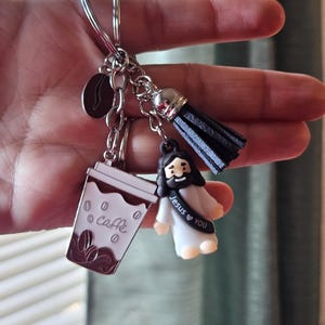 May include: A silver keychain with multiple charms. Charms include a coffee cup with "Caffe" text, a coffee bean, a black tassel, and a small Jesus figure with "Jesus &hearts; YOU" text. The coffee cup is white and brown.
