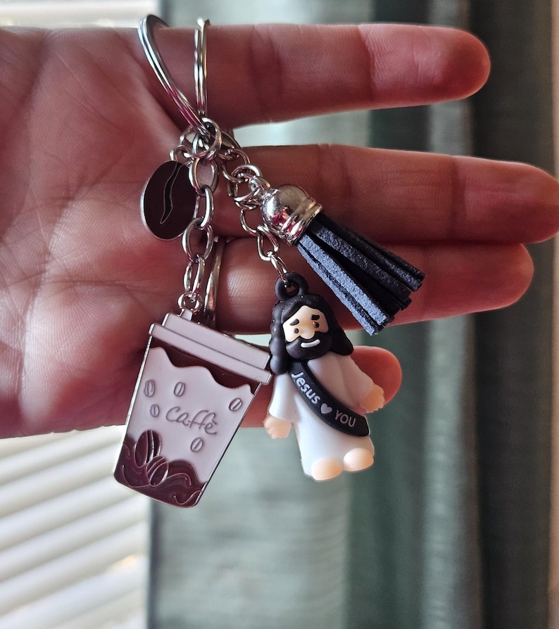 May include: A silver keychain with multiple charms. Charms include a coffee cup with "Caffe" text, a coffee bean, a black tassel, and a small Jesus figure with "Jesus &hearts; YOU" text. The coffee cup is white and brown.