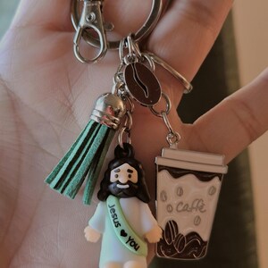 May include: A silver keychain with a green tassel, a coffee bean charm, and two decorative charms. One charm is a small figure of Jesus with the text "Jesus &hearts; You". The other is a coffee cup with the word "Caff&egrave;".