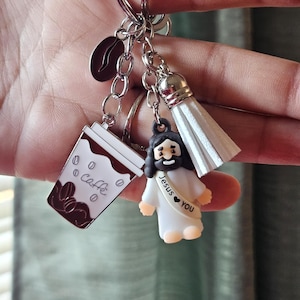 May include: A silver keychain with multiple charms. Charms include a coffee cup with "Caff&egrave;" text, a coffee bean, a white tassel, and a small Jesus figure with "Jesus ❤️ You" text. The charms are attached to a silver keyring.