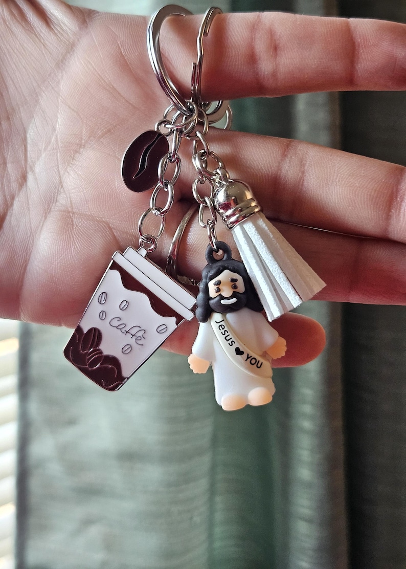 May include: A silver keychain with multiple charms. Charms include a coffee cup with "Caff&egrave;" text, a coffee bean, a white tassel, and a small Jesus figure with "Jesus ❤️ You" text. The charms are attached to a silver keyring.