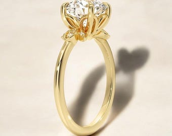 Könnte beinhalten: Ein goldener Ring mit einem großen, klaren, runden Edelstein, der in einem zarten Blumenmuster gefasst ist. Der Ring hat ein schlichtes Band.