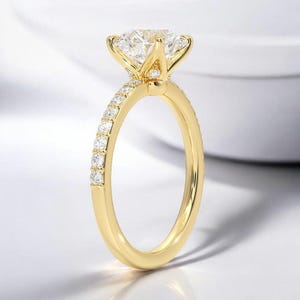 Puede incluir: Un anillo de compromiso de oro con una piedra central grande, redonda y transparente y una banda de piedras más pequeñas y transparentes.