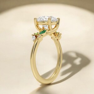 Puede incluir: Un anillo de oro con una piedra central grande, redonda y transparente y piedras más pequeñas, verdes y transparentes, engastadas en un delicado diseño de hojas.