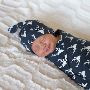 May include: A newborn baby wearing a navy blue and white deer print knotted hat and swaddled in a matching navy blue and white deer print swaddle blanket.