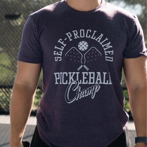 May include: A man wearing a purple t-shirt with the text "Self-Proclaimed Pickleball Champ" in white. The shirt features a graphic of two pickleball paddles and a ball. He is holding a yellow pickleball.