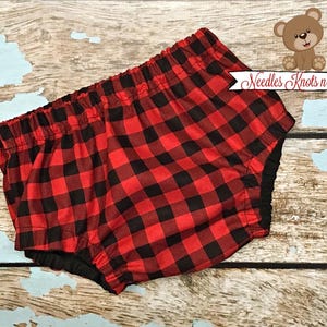 May include: Red and black plaid baby bloomers with a black elastic waistband. The bloomers are made of a soft, comfortable fabric and have a ruffled edge.