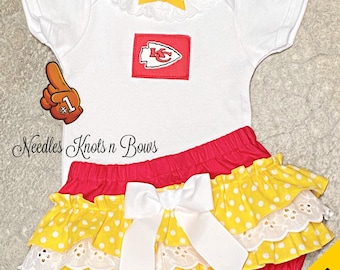 May include: A white baby bodysuit with a Kansas City Chiefs logo patch, a yellow bow, and red and yellow ruffled shorts. The shorts have a white bow and a polka dot pattern. The image also includes a foam finger.