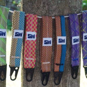 Może przedstawiać: Kolekcja kolorowych, tkanych pasków z czarnymi metalowymi klipsami, zawieszonych na białej linie. Każdy pasek posiada białą etykietę z niebieskimi literami "SW". Paski prezentują różne wzory i kolory, w tym zielony, beżowy, różowy, pomarańczowy, niebieski i fioletowy.