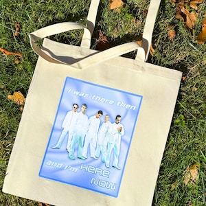 Puede incluir: Bolso tote de color crema con un gráfico azul que muestra un grupo de personas con trajes blancos. El gráfico dice "I was there then" y "and I'm HERE NOW". El bolso tiene dos asas color crema y está sobre hierba verde.