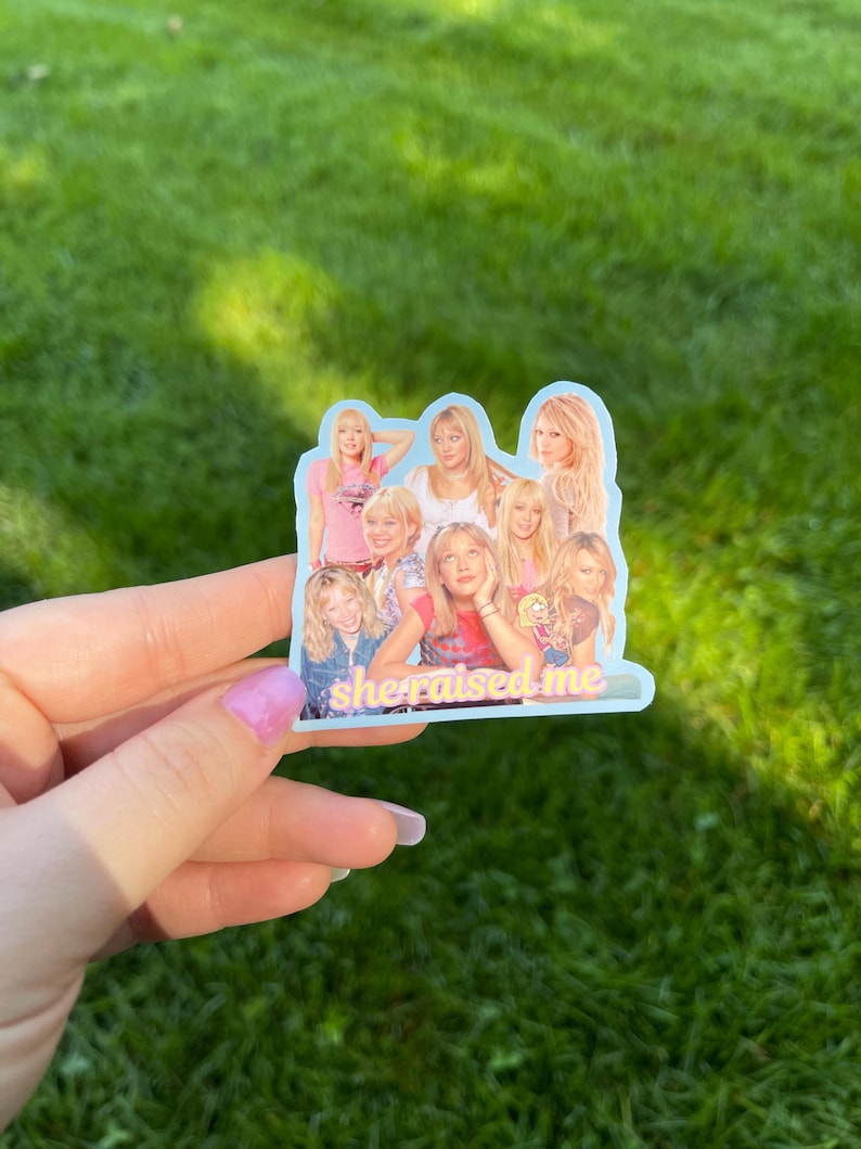 May include: A sticker featuring a collage of images of women, with the text "she raised me" in a script font. The sticker has a light blue border and is held against a background of green grass.