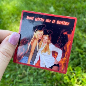 May include: A rectangular sticker with a red border and the text "bad girls do it better." The sticker features a photo of two women with bunny ears, set against a fiery background.