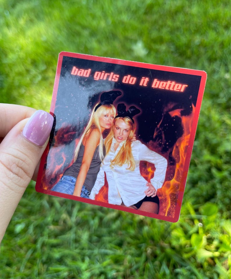 May include: A rectangular sticker with a red border and the text "bad girls do it better." The sticker features a photo of two women with bunny ears, set against a fiery background.