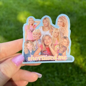 May include: A light blue sticker featuring a collage of images of a woman. The sticker has the text "she raised me" in a pink and yellow font. The background is green.