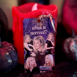 May include: A rectangular keychain with a collage of images and the text "what is normal?" in white. The keychain has a silver-coloured chain and is set against a blurred background with a red candle and decorative pumpkins.