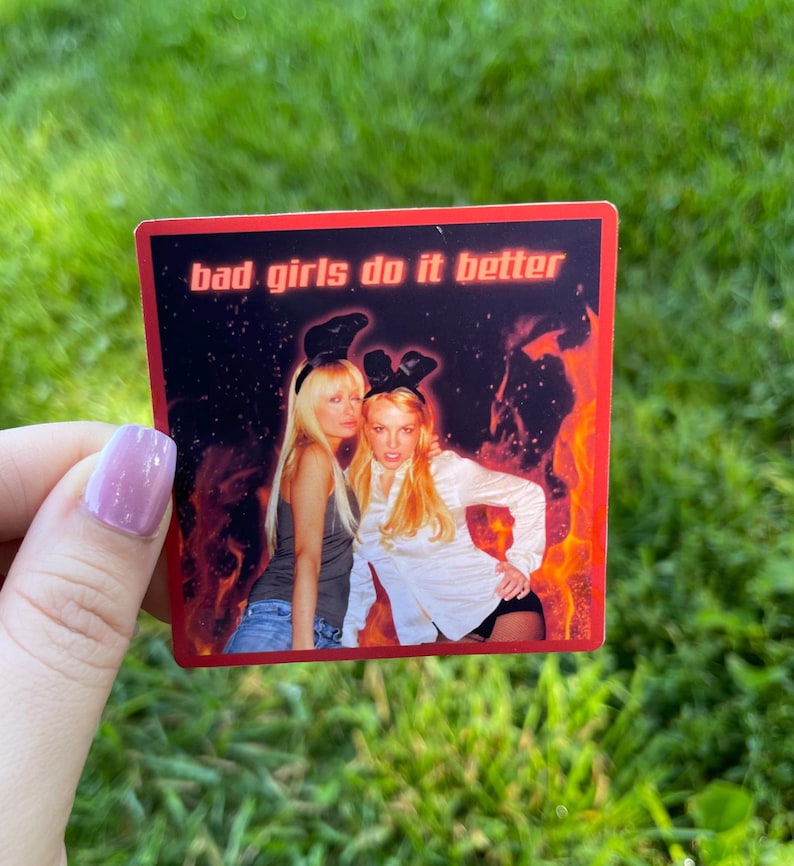 May include: A square sticker with a red border and the text "bad girls do it better" on a black background. The sticker features two women in front of a fiery background. One woman wears a gray top and jeans, the other a white shirt.