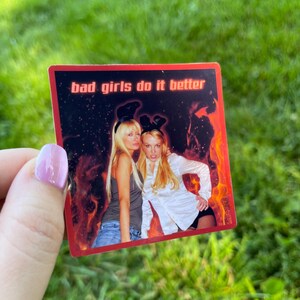 May include: A rectangular sticker with the text "bad girls do it better" in white. The sticker features two women with long blonde hair, one wearing a gray tank top and the other a white button-down shirt. The background has a red and orange flame design.