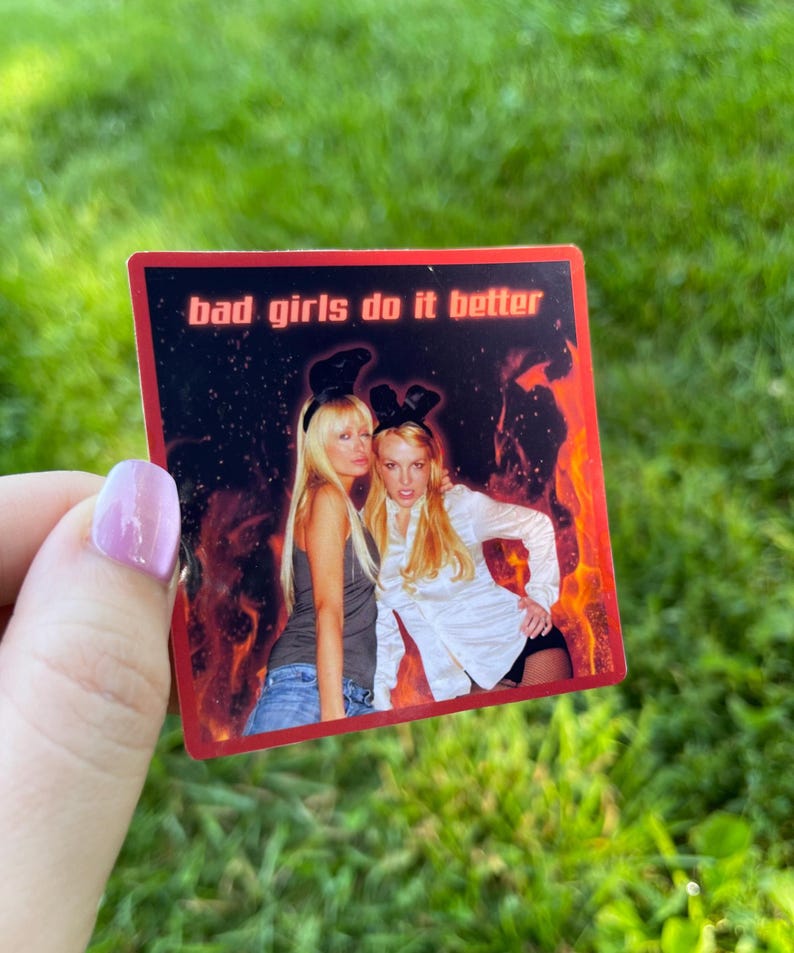 May include: A rectangular sticker with the text "bad girls do it better" in white. The sticker features two women with long blonde hair, one wearing a gray tank top and the other a white button-down shirt. The background has a red and orange flame design.