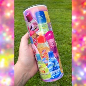 May include: A tumbler featuring a collage of colorful body spray bottles. The bottles are various shapes and sizes, with labels in different colors and fonts. The tumbler has a glossy finish and is held against a background of green grass.