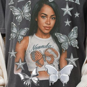 May include: Dark grey t-shirt with the name "Aaliyah" in silver lettering and a portrait of a woman. The shirt features butterfly and star graphics. The woman in the portrait wears a white tank top with a butterfly design and the name "Naanda".