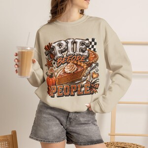 May include: A beige crewneck sweatshirt featuring a graphic print of a slice of pumpkin pie with whipped cream, a whole pie, a pumpkin, and cinnamon sticks. The text reads PIE BEFORE PEOPLE in bold, stylized lettering with a checkered pattern accent.