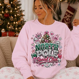 Op de afbeelding: Een lichtroze sweater met ronde hals en een grafische print van een versierde kerstboom, discoballen, cadeautjes en zuurstokken. De tekst luidt North Pole Social Club in opvallende, metallic-achtige letters met diverse kersticonen.
