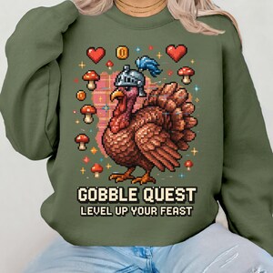 May include: Olive green sweatshirt featuring a pixelated turkey wearing a knight's helmet, surrounded by hearts, mushrooms, and coins. The text "GOBBLE QUEST LEVEL UP YOUR FEAST" is displayed below the turkey. A fun, festive design.