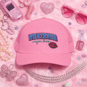 May include: Pink trucker hat with the word "WHATEVER" embroidered in blue and red, along with "major loser" and a red lip graphic. The hat is surrounded by pink accessories, including sunglasses, a phone, and jewelry.