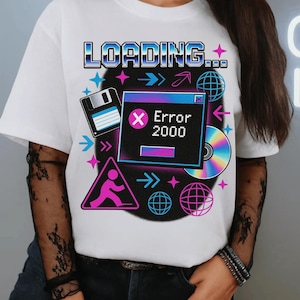 May include: White t-shirt with a retro computer-themed graphic. The design features the word "LOADING..." in a blue and black pixelated font, along with a computer error message, a floppy disk, and a CD.
