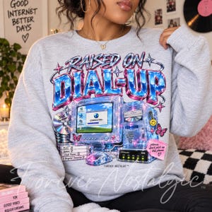 May include: A light gray crewneck sweatshirt featuring a graphic print with the text RAISED ON DIAL-UP. The design includes a vintage computer, a CD, a mouse, and a pink sticky note with the text please don't pick up the phone.