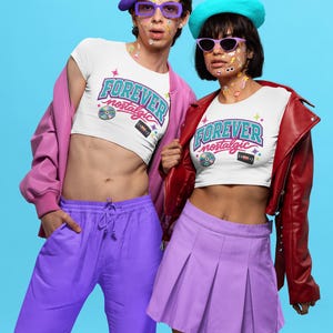 Könnte beinhalten: Zwei Personen tragen weiße Crop-Tops mit dem Text "FOREVER nostalgic" in Retro-Schrift. Eine Person trägt eine lila Hose und eine lila Baskenmütze, die andere einen lila Rock und eine blaue Baskenmütze. Beide tragen Sonnenbrillen und haben Aufkleber im Gesicht.