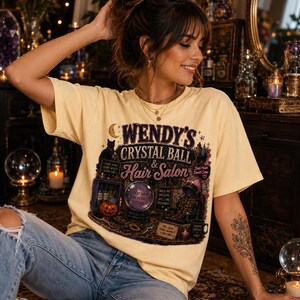 Op de afbeelding: Een lichtgeel T-shirt met korte mouwen en een grafische print met de tekst Wendy's Crystal Ball & Hair Salon. Het ontwerp bevat een zwarte kat, een glazen bol, een pompoen, een kappersstoel en diverse mystieke en haarstylingaccessoires.
