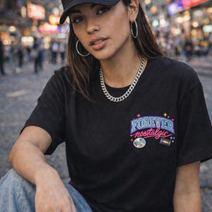 May include: Black baseball cap and t-shirt with the text "FOREVER nostalgic" in a colorful design. The cap has a curved brim and the t-shirt has a small graphic on the front. The person is wearing silver hoop earrings and a silver chain.